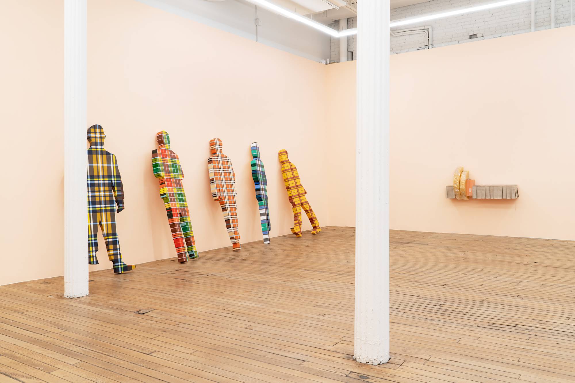 Five flat, life-sized human figures covered in plaid patterns lean against a peach wall in an art gallery. On the right wall, a shelf lined with a short, beige, pleated curtain holds two yellow cushions and an orange shoe box.