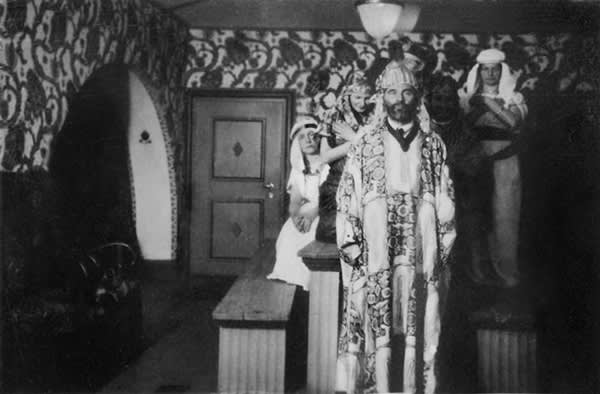 Black and white vintage photo showing several people in costumes engaged in an occult ritual in a room with pattered wall paper.