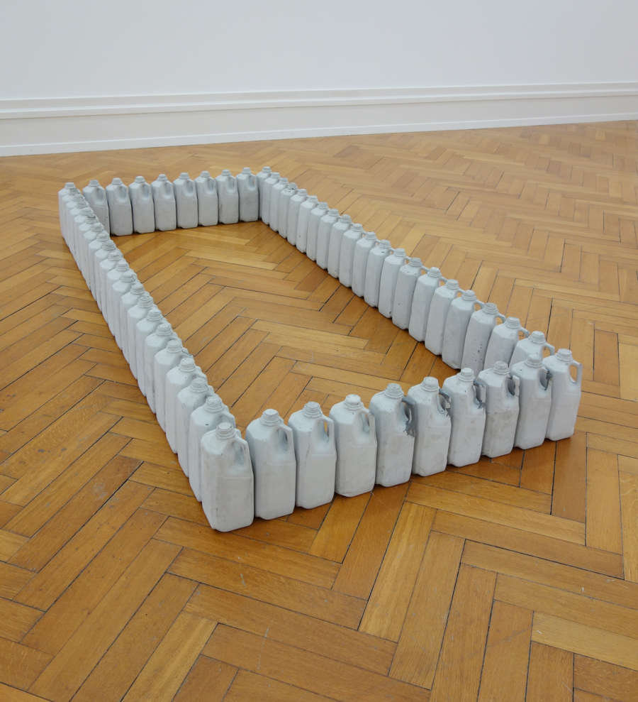 In a gallery space, numerous milk jugs cast in concrete arranged in the rectangular form of a grave plot on the floor. 