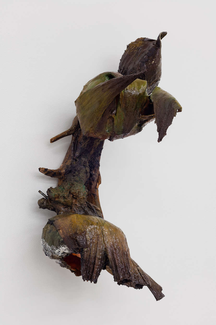 Image of a sculpture by Brandon Ndife. Hanging on a gallery wall is a brown/black plant like growth covered in dirt and debris. There are cavities created by corn husks that hold some other objects, all clinging to a few sticks and branches.