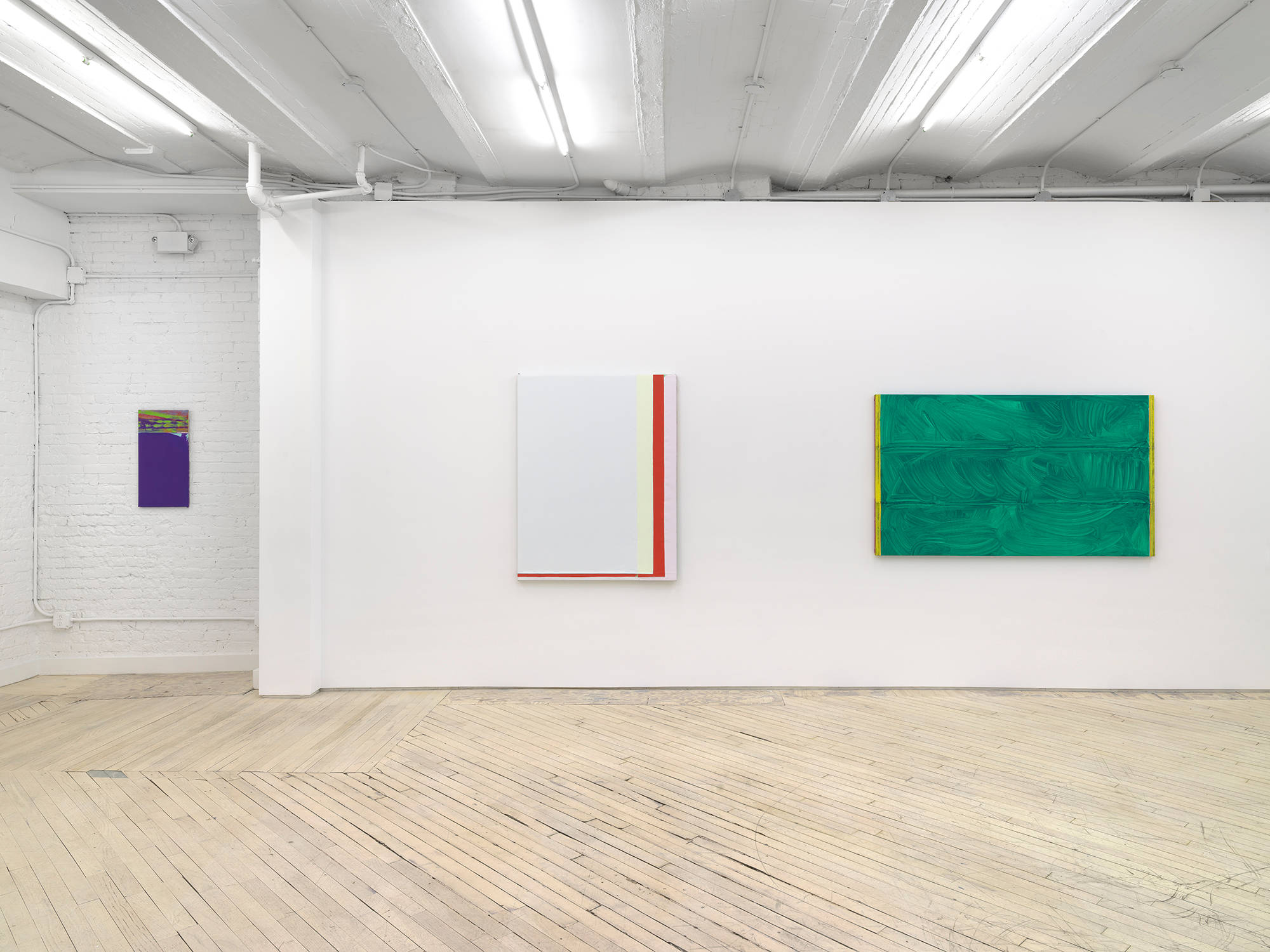 Along a gallery wall. A thin purple painting hangs to the left. In the middle is a larger white monochrome painting with a red band of color on the right and bottom sides. To the right a horizontal green painting with a brushy surface.