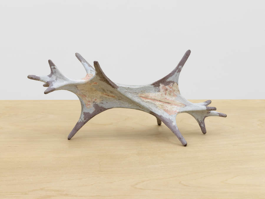 On a wooden table, a small abstract sculpture resembling an organic form, shell, or bone. The piece is gray with pinkish accents. 
