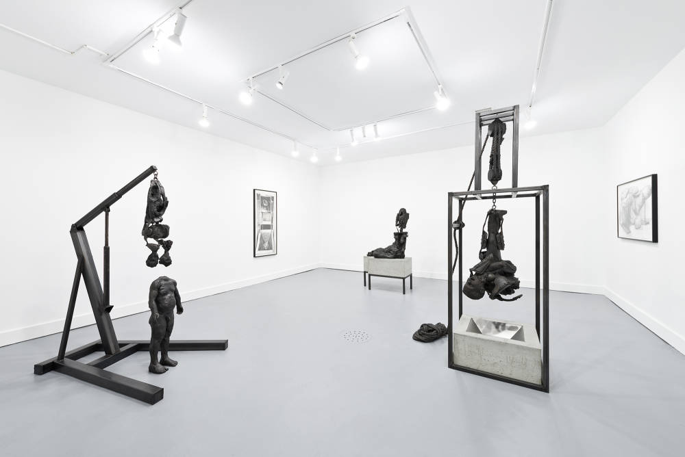 Installation view of three metal and concrete sculptures, mostly depicting decaying figures, with two framed drawings on the walls behind them.