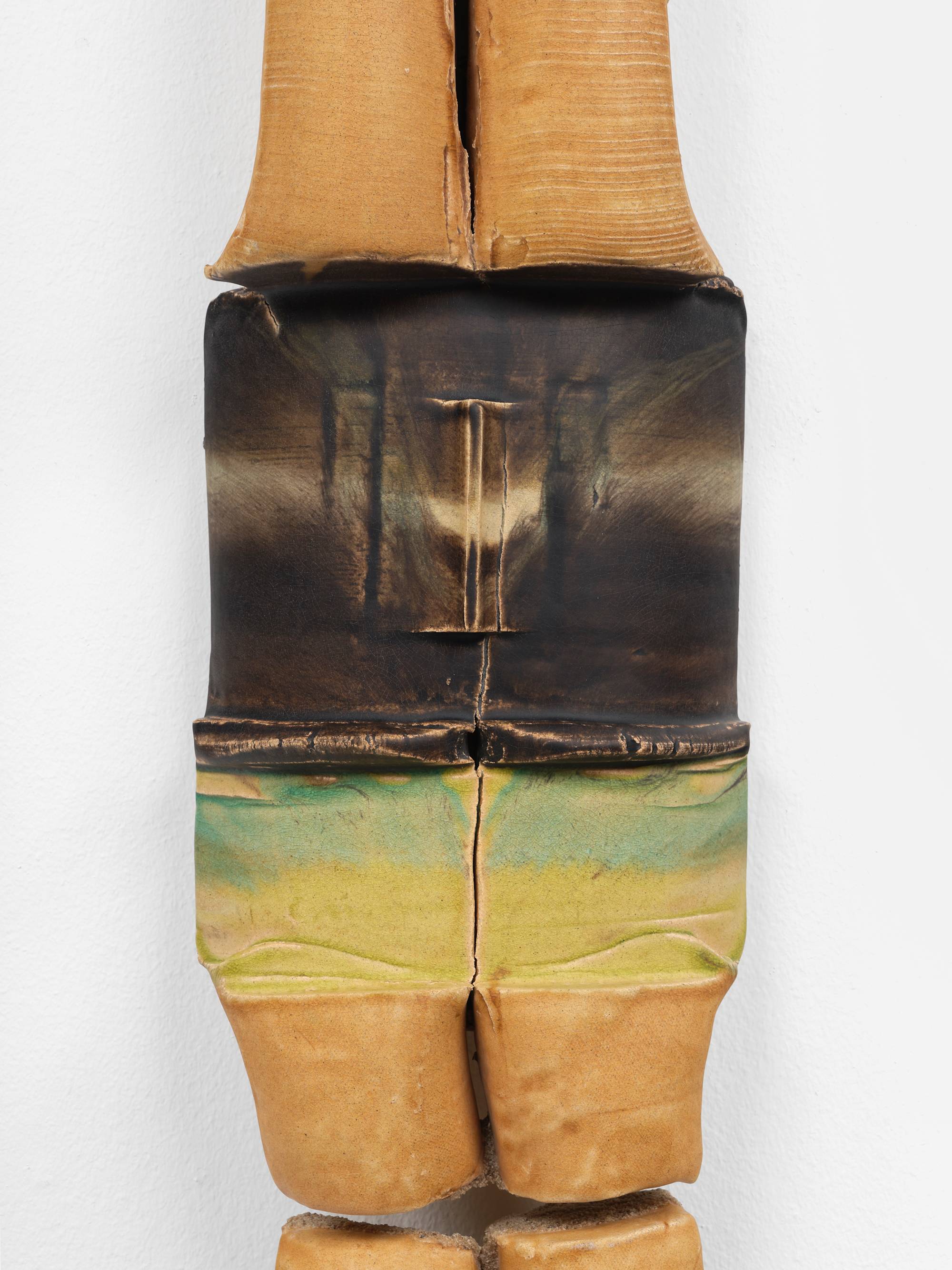 Detail of a large, vertically oriented, 2-piece sculpture, featuring cylindrical elements, in hues of mustard, brown, and green, hanging on a white wall.
