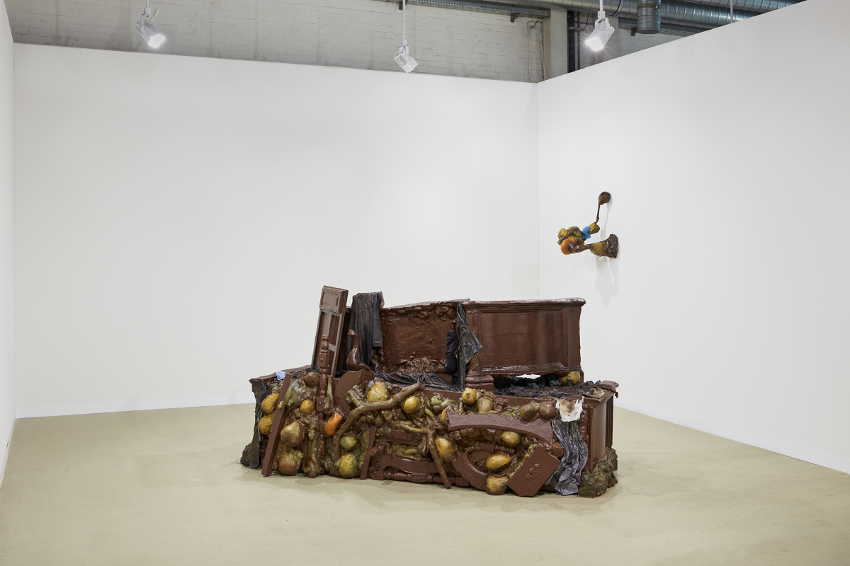 A large piece of brown furniture sits in the middle of a room with white walls. A smaller sculpture hangs to the right of the larger installation on the wall.
