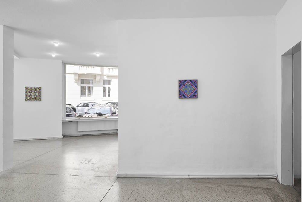 Image of a gallery space with a wall on the right, with a painting installed. To the left, there is a father wall with another painting installed. There is a window in the background as well. 