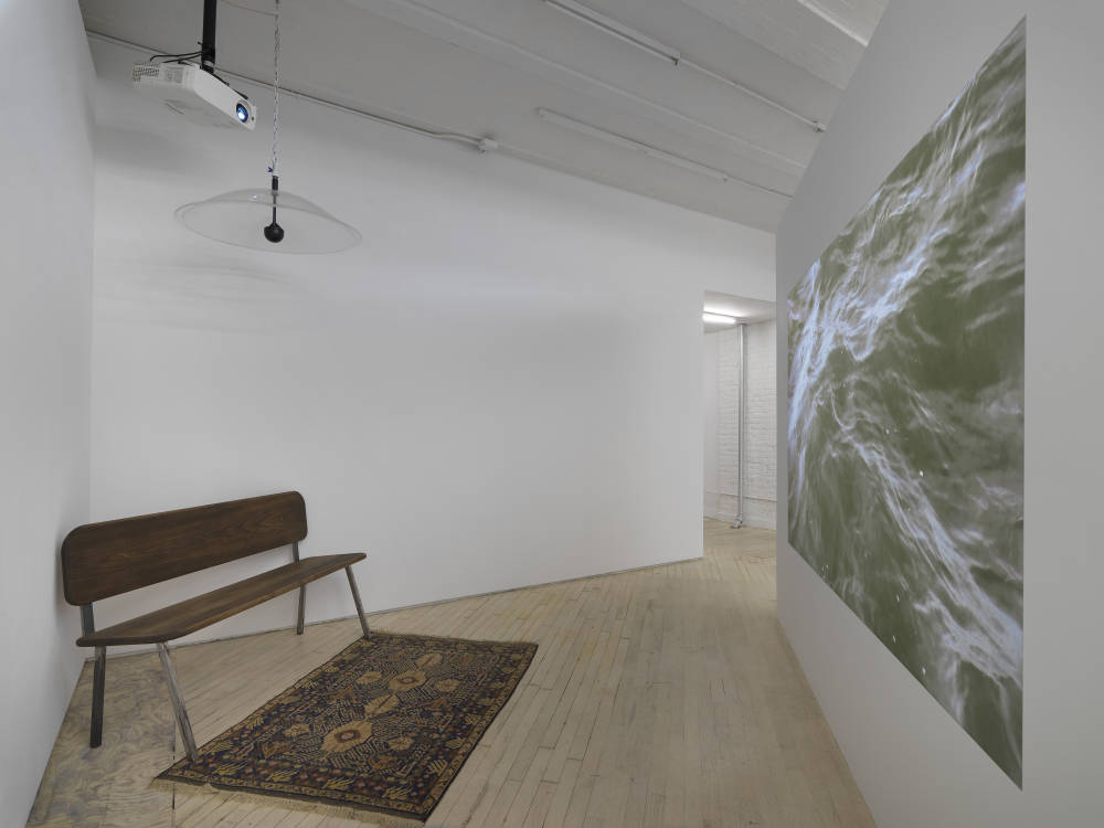 In a corner of a gallery space, a large digital projection of moving water with a bench and rug. 