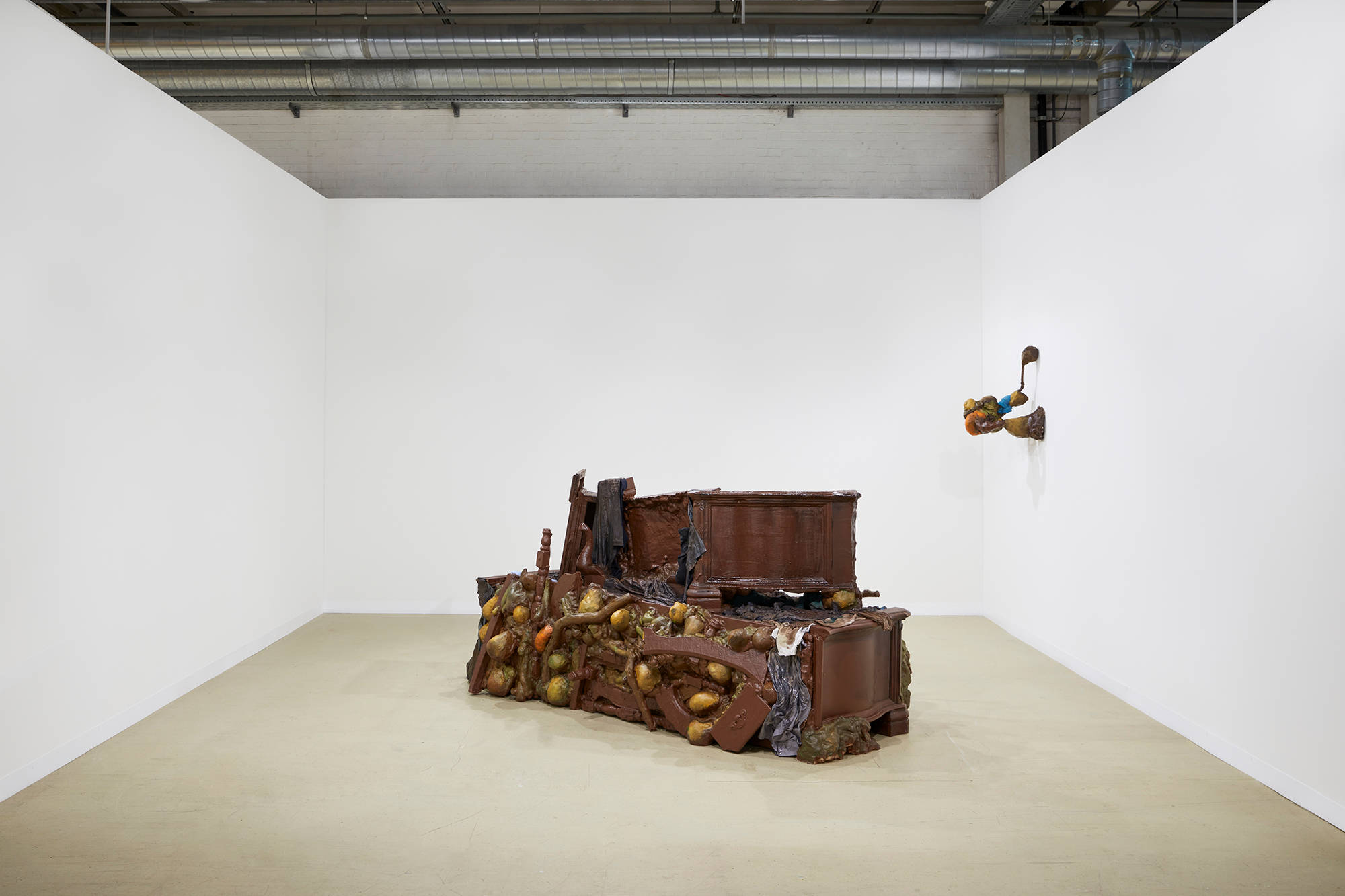 A large piece of brown furniture sits in the middle of a room with white walls. A smaller sculpture hangs to the right of the larger installation on the wall.