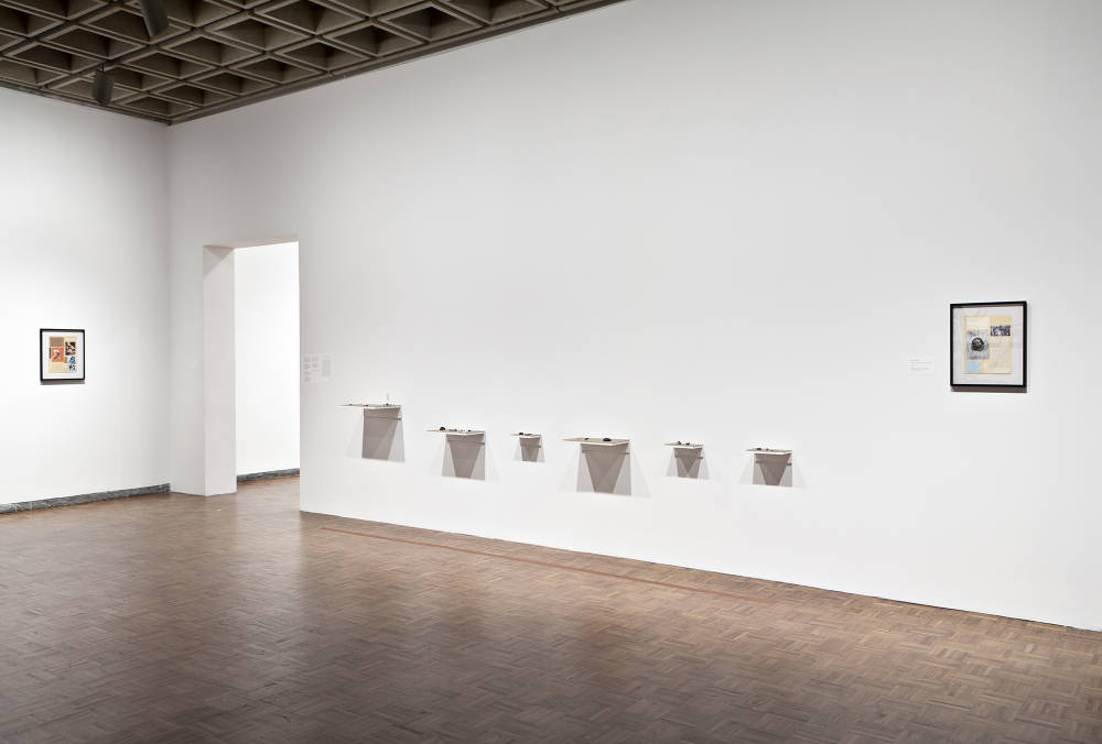 In a large exhibition space in a museum, numerous wall mounted shelves are hung in aline with sculptures on top of them. To the left and right of the sculptures are artworks in black frames. 
