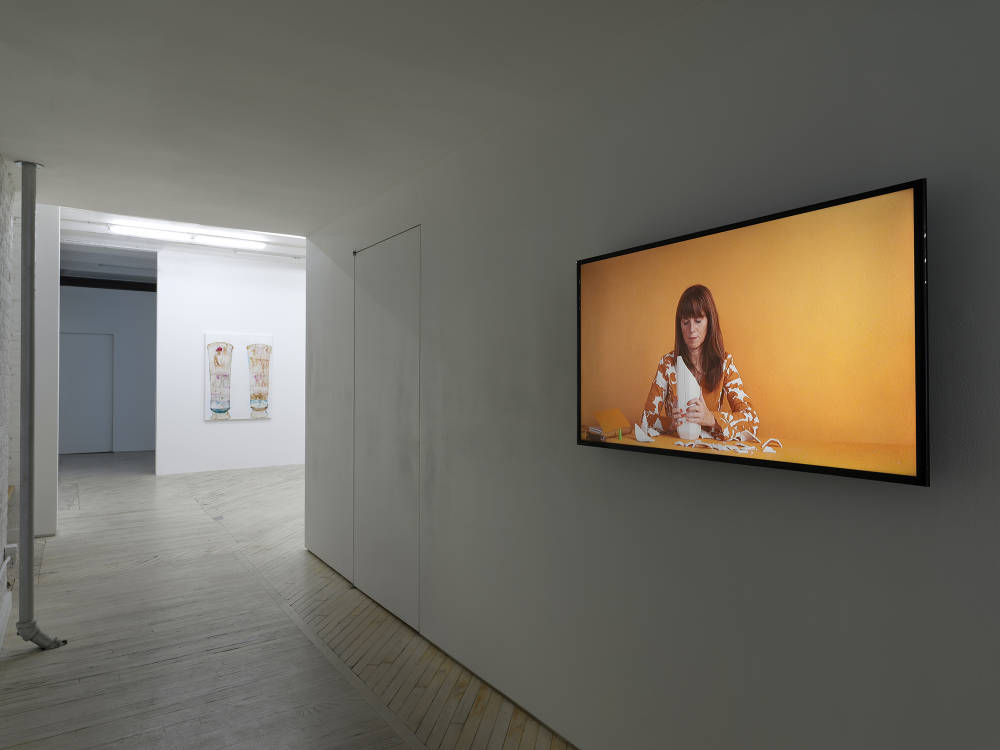 Installation view of a dark hallway in the gallery, on the right wall is a video of a woman sitting in front of an orange background. At the distance, beyond the hallway, is a painting hanging on the wall of two ancient vases.