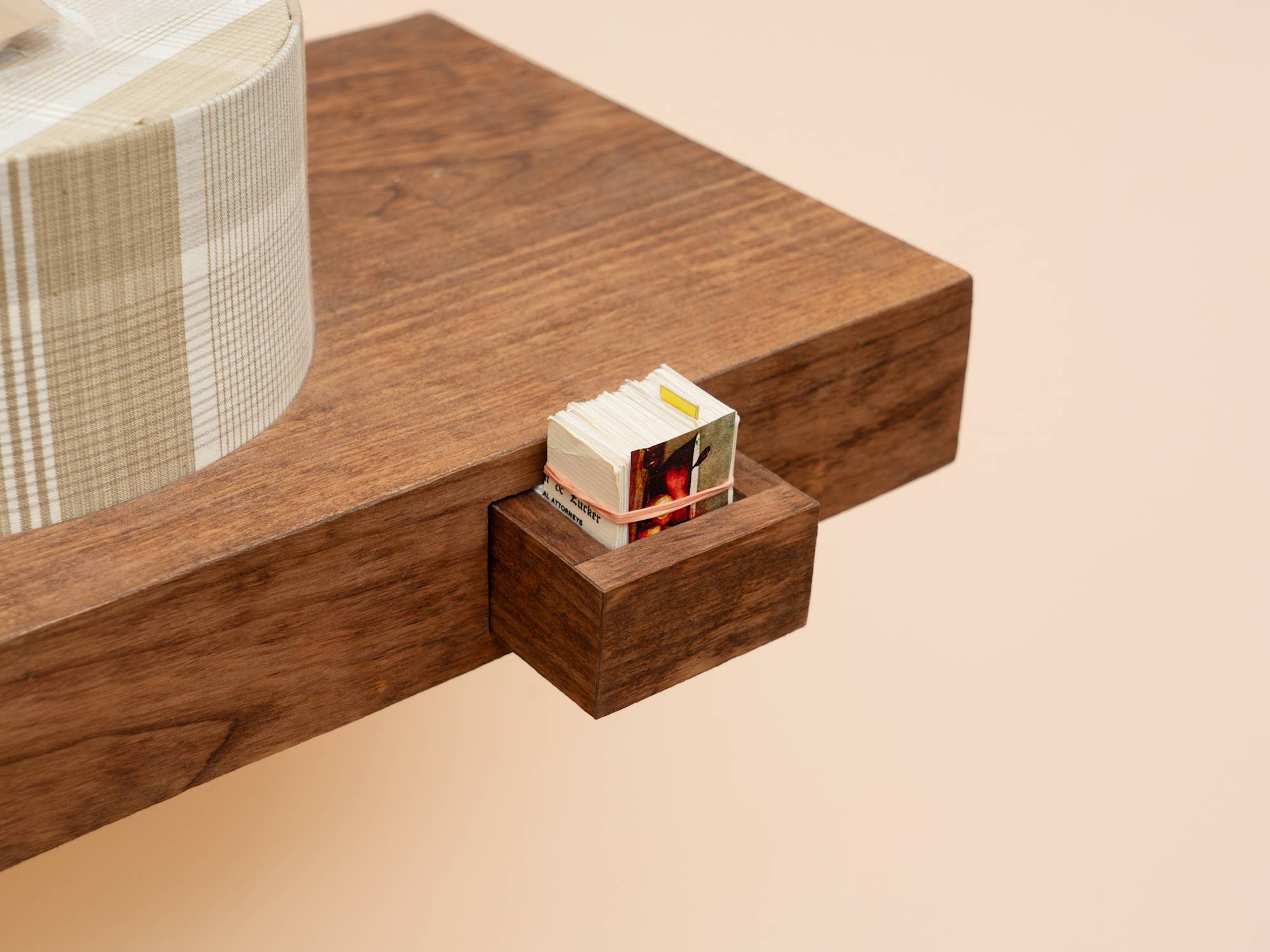 Detail of a wood veneered shelf hangs on a peach colored wall. 2 tan, plaid, circular gift boxes sit on the shelf with tan socks in plastic bags draped over them. A small drawer juts out of the front of the shelf and is neatly filled with trimmed business cards. A large, fake audio cassette case featuring a truck driving over a bridge hangs above the shelf, wrapped in cellophane and decorated with three silver gift bows.