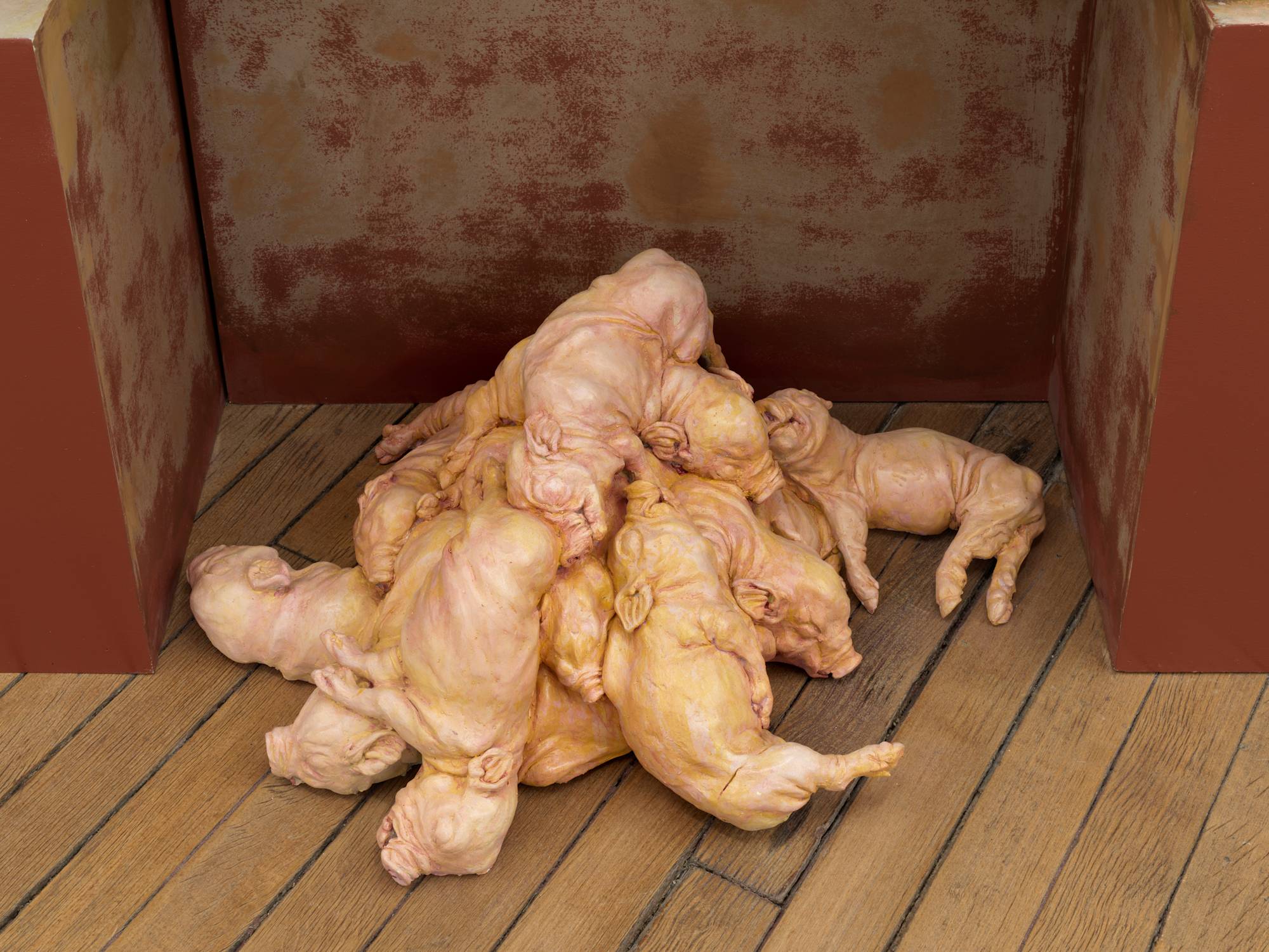 A sculpture depicting a pile of piglets resting on a wooden floor with red paint behind them on a wooden structure.
