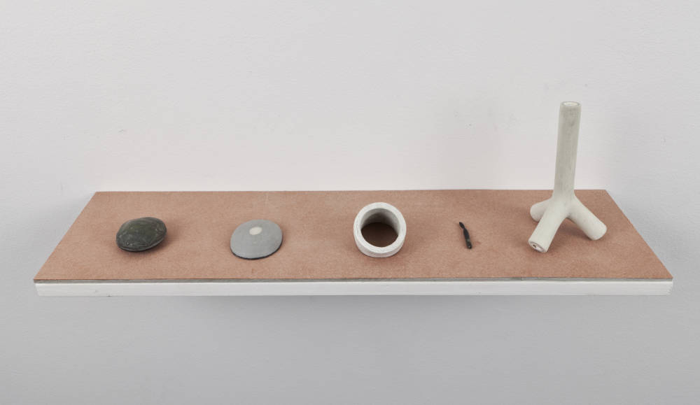 An orange wall-mounted shelf attached to a white wall. Spaced evenly apart are miniature sculptures resembling bone forms or rocks.