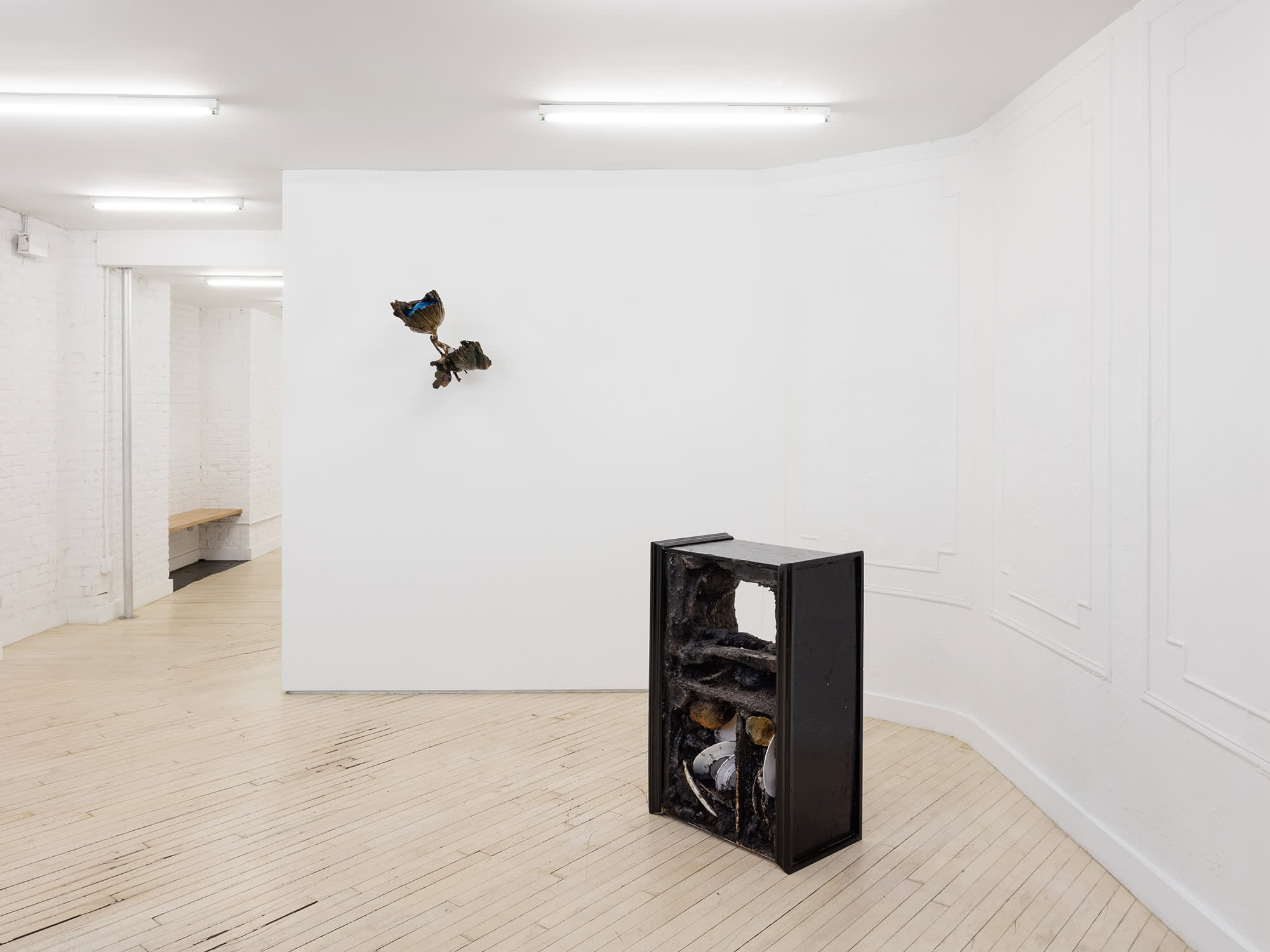 Installation view of an exhibition by Brandon Ndife. In a gallery there are two sculptures, one hanging on the wall and one in front of it on the floor. Both are brown/black and covered in dirt and debris. The wall sculpture appears like a plant and a sconce. The floor sculpture looks like a piece of furniture.