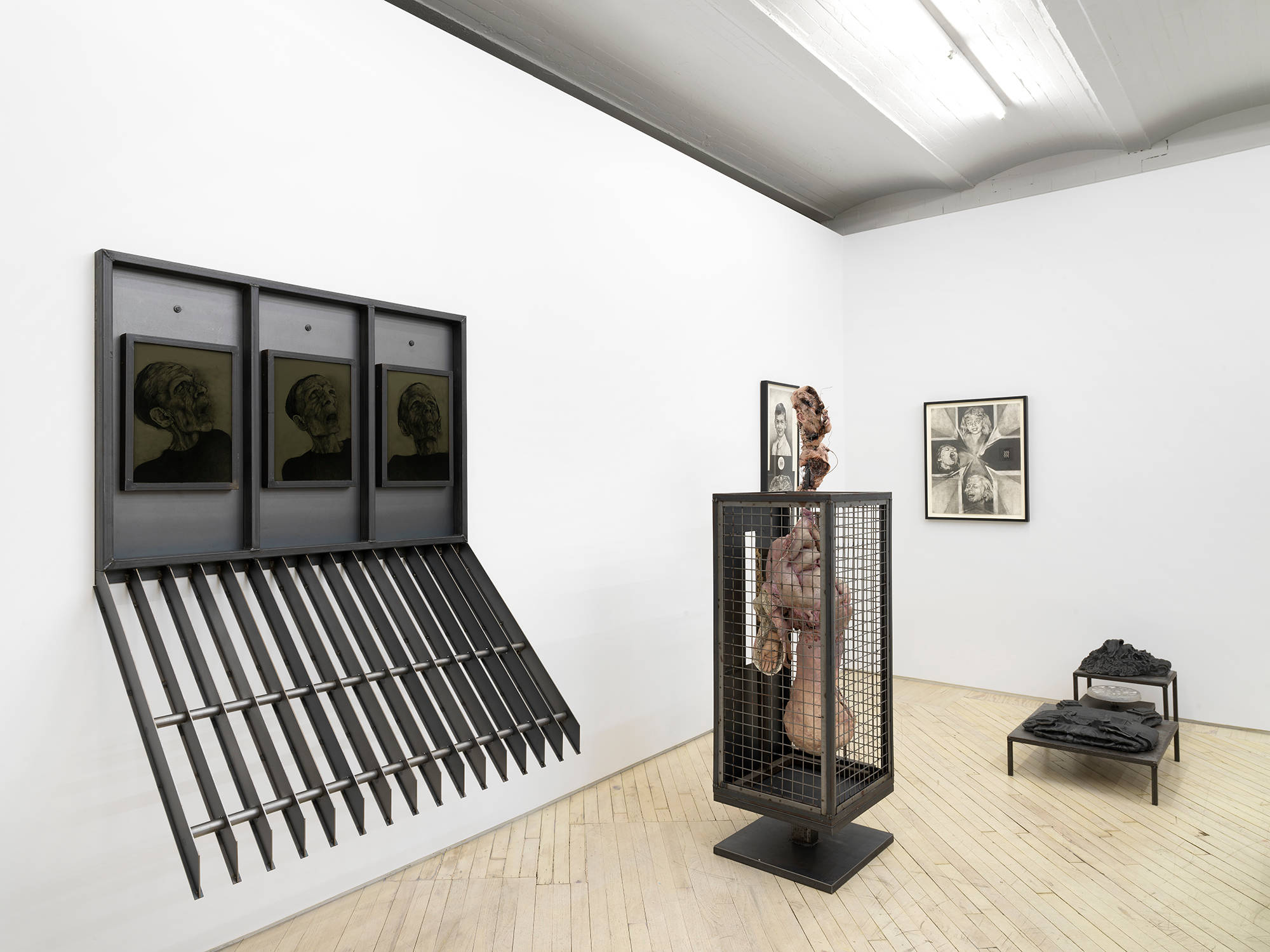 A gallery space filled with large metal sculptures resembling cages. Hanging on the wall are two framed graphite drawings.