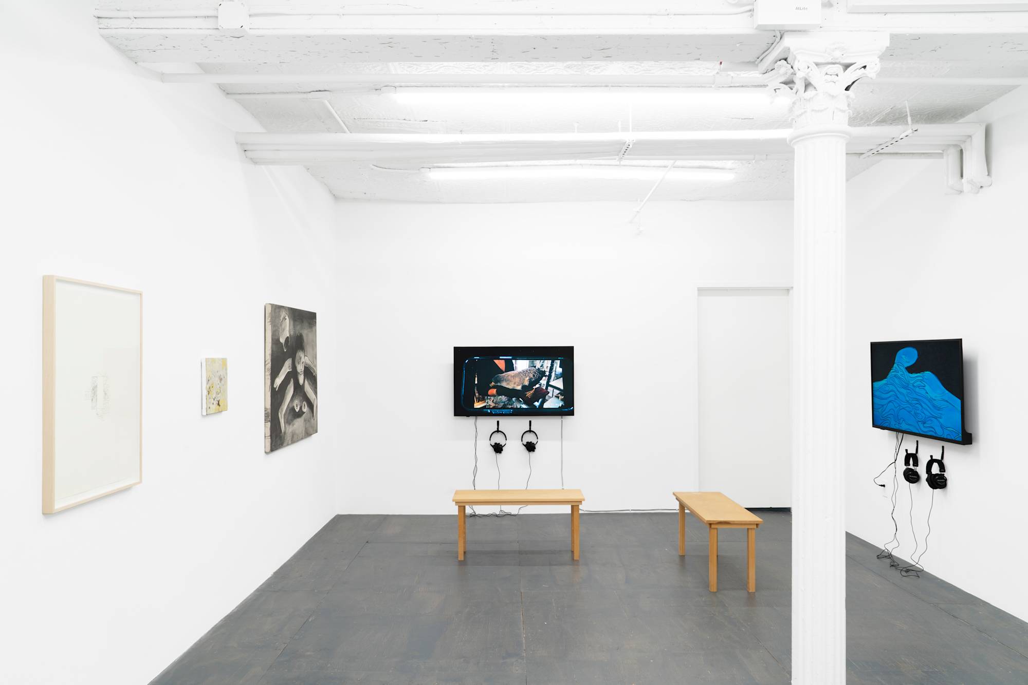 A view of a basement gallery with grey floor and white column. Two paintings, one framed graphite drawing, and two films displayed on monitors hang on the walls. The monitors are accompanied by headphones and wood benches.