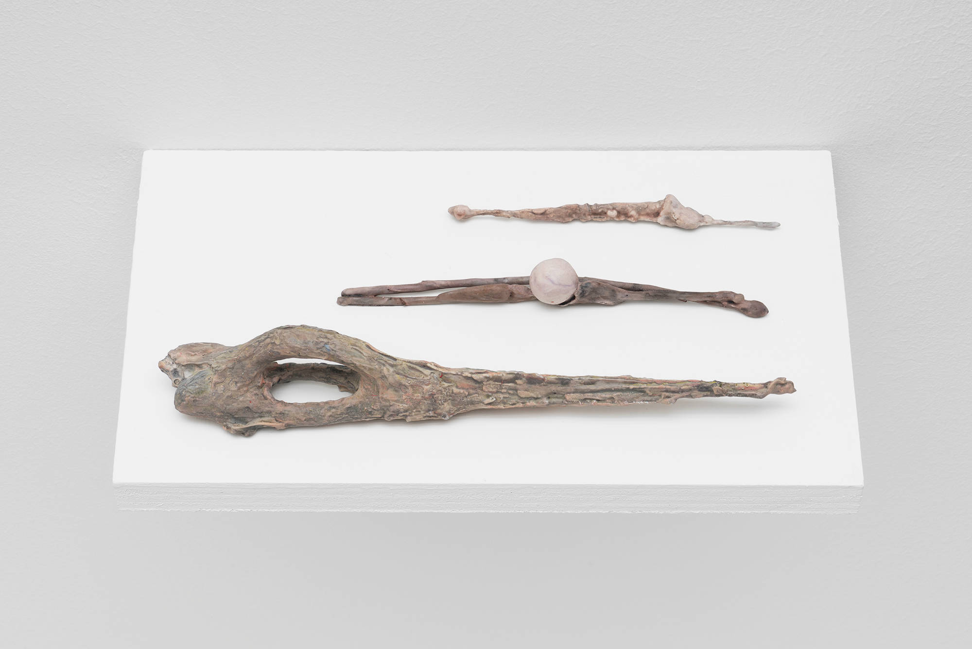 A trio of small, handmade sculptures by Matt Hoyt sit on a white ply wood shelf. They are light brown and pink in color and resemble wands.