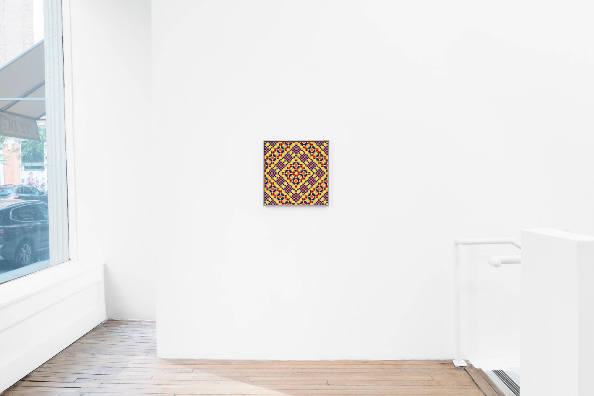 A geometric abstract artwork featuring a repeating pattern of squares and diamonds in shades of red, yellow, and purple hangs in a bright lit gallery. A stairwell descends to the right, out of frame. A view of the street is visible from the window to the left of the painting.