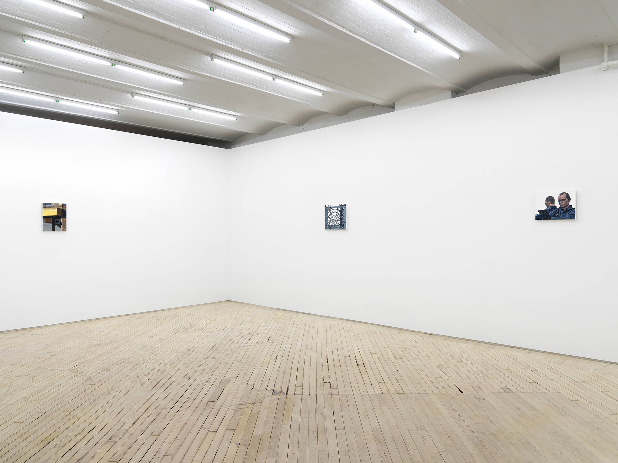 Angled view of the gallery with white walls, fluorescent lights and light wood floors. Three very small paintings hang on the 2 visible walls. At left is a color painting showing a yellow awning on a city street and at right are two paintings, a grey and white square painting of a grey milk crate, and a portrait of a man with glasses who is holding a gun.