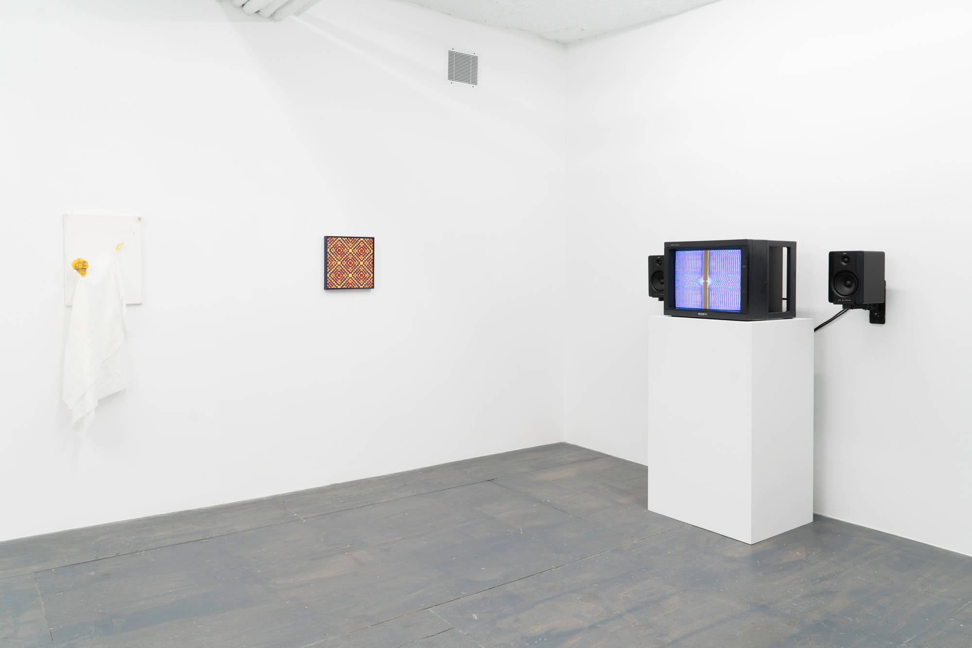 A white painting with a puppet-figure adhered to it hangs next to a geometric painting consisting of a pattern of repeating diamonds and triangles hung along a gallery wall. To the right a tv monitor displaying a still image blurred colors in a grid pattern taken from an animation, resting on top of a white pedestal with black speakers mounted to the wall on either side.