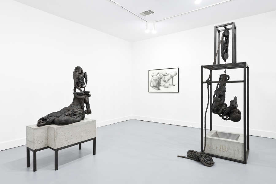 Installation view of two metal and concrete sculptures, mostly depicting decaying figures, with a framed drawing on the walls. 