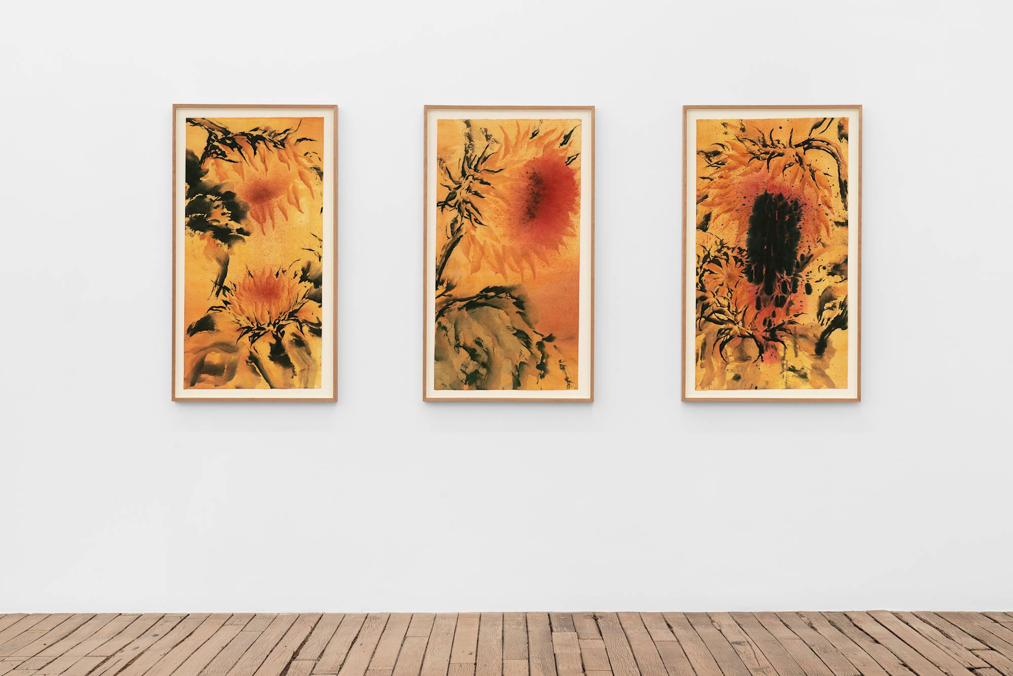 Three vibrant ink paintings on Japanese paper featuring abstracted sunflowers against an orange background in a wood frames hang in a row about a wood floor in a gallery.