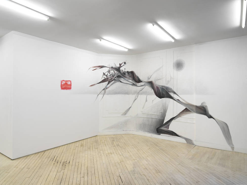 In a gallery space, a small photograph of a building printed in red, to the right is a large wall drawing of an abstracted form crawling toward the photograph.