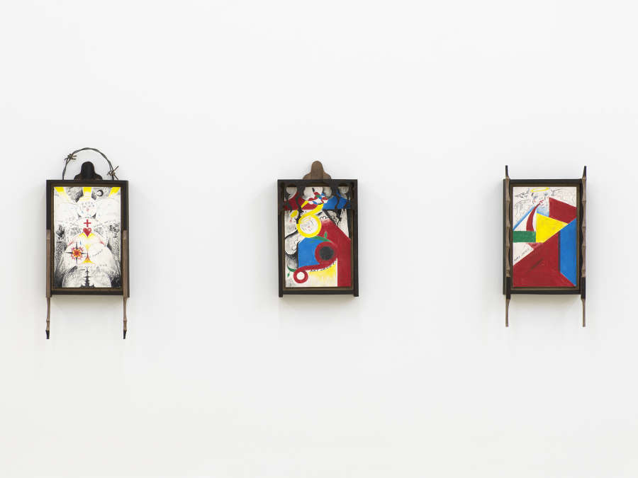 Installation view of three small vertical format drawings hanging on the wall all are framed in black wood with ornately carved frames. The first from the left also has barbed wire in an arc along the top edge of the frame. The drawing is black and white with accents of yellow and red, and shows concentric circles arranged geometrically, with rays of yellow sun at the top, symbols throughout, such as a red cross and red heart at center and a crescent moon and angel face which has the word 'Joy' written across the forehead.  At left and right are two brick smokestacks. The two drawings on the right have solid irregular but geometric shapes in yellow, red, blue and green, as well as cartoonish angel heads with wings and haloes and the words 'Art Saves!' written in pencil.