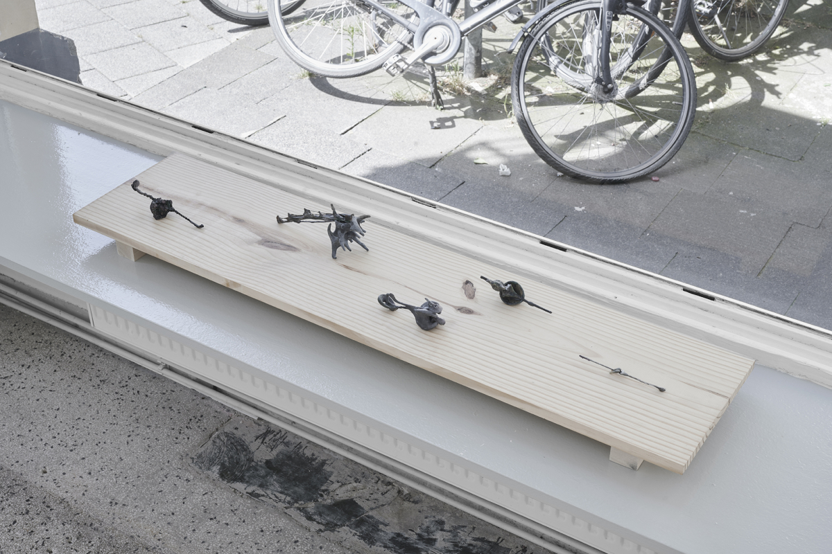 Five small hand-sized sculptures are presented on a rectangular piece of wood in front of a large window that opens up to a street view. The sculptures are amorphous, skeletal, and gray. They are resting on a bench inserted between two walls. 