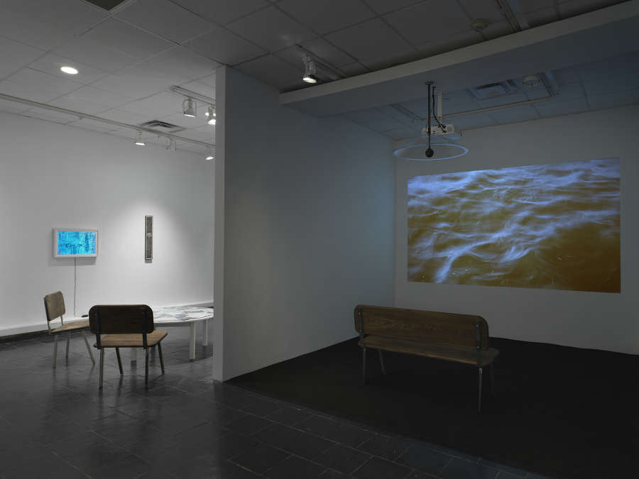 A projection of a body of water with a bench in front of it. In the background a small round table containing ephemera. A blue lightbox is hung on a wall in the distance.