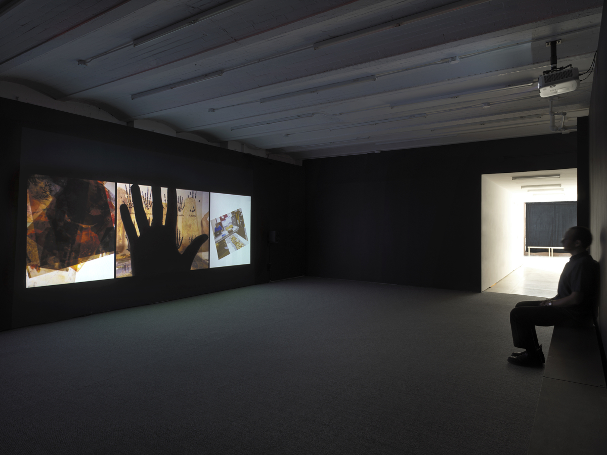 View of Gallery space with Video Installation projected onto dark wall while man sitting across the room watches. The still image contains a hand in the middle. 