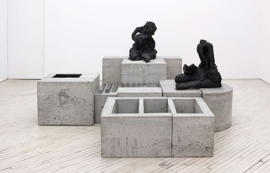 Image of a concrete and cast iron sculpture with blocks of varying heights and two decaying figures siting adjacent each other.