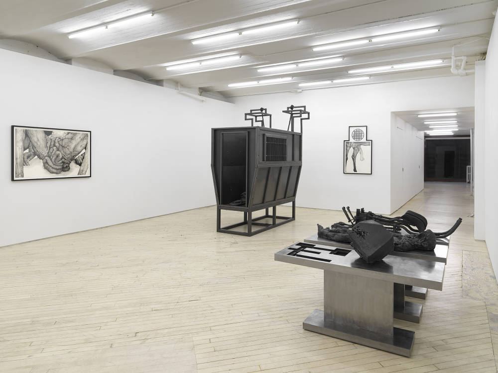 In a gallery space, a large metal and cast iron sculpture made up of different components. Resting on the base are abstract remnants of bodies next to a cube form. In the center of the gallery is a large metal sculpture resembling a cattle container. Behind the sculptures on the walls are two graphite drawings of genitals in black frames. 