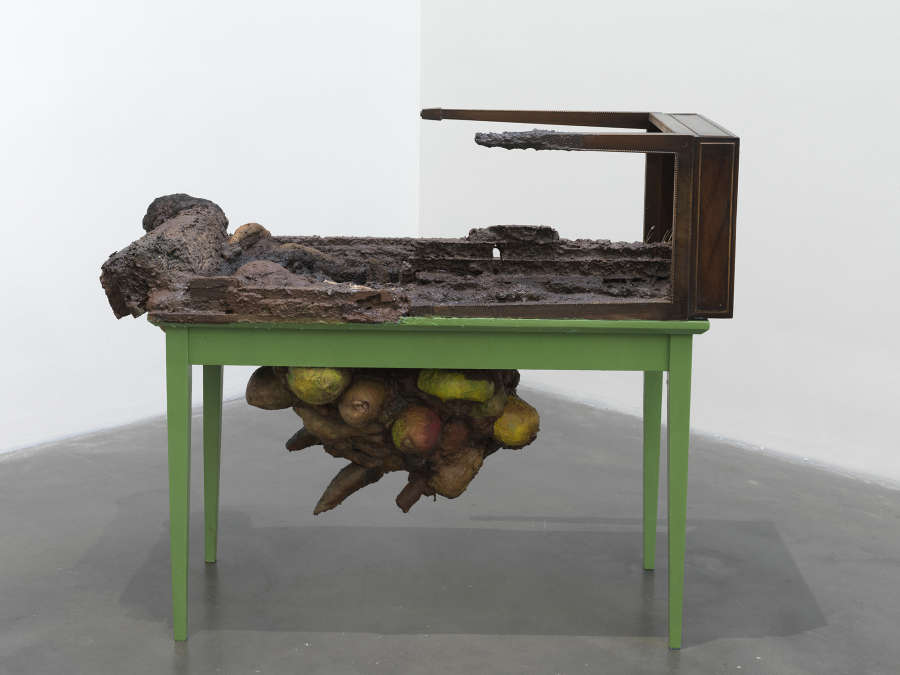 A free standing sculpture resembling a table with additional legs attached to the top. In the center is a large form growing out of the sculpture that resembles fungi or other vegetation. 