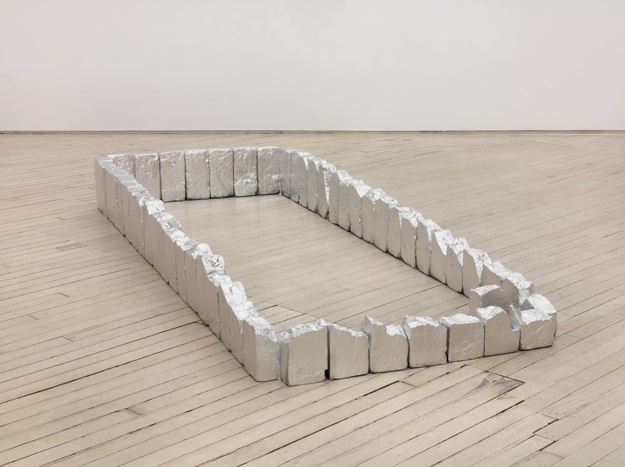 Image of Tom Holmes sculpture installed on the floor of a gallery which appears to be a grave plot demarcated by a series of bricks that are coated in aluminum foil.