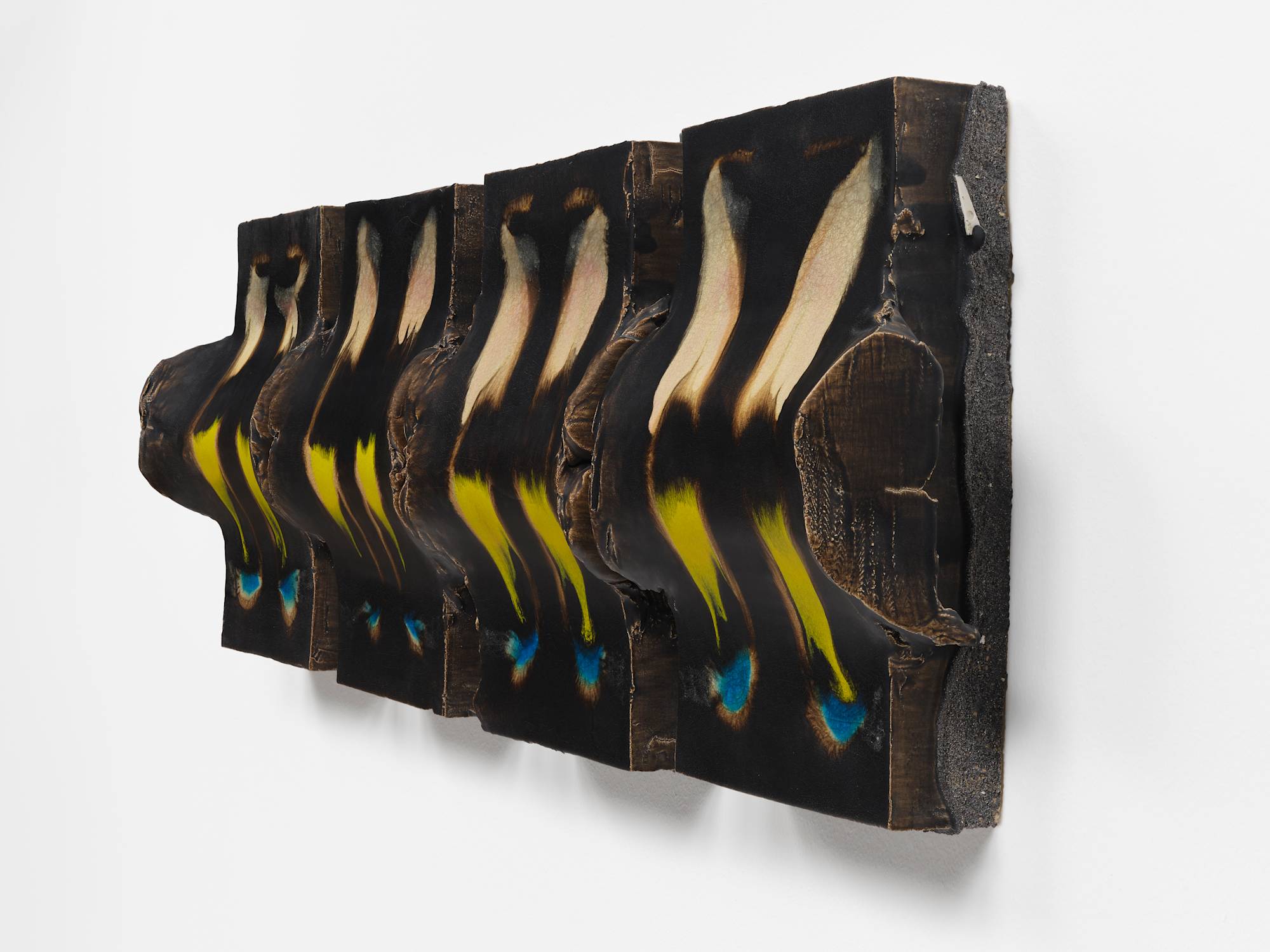 Three quarter view of four nearly identical vertical cuboid ceramic sculptures hang in a tight horizontal line on a white wall. Each piece has two horizontal cylindrical bands dividing their composition beneath the smooth top surface that has been glazed dark brown with accents of tan, green, and blue.