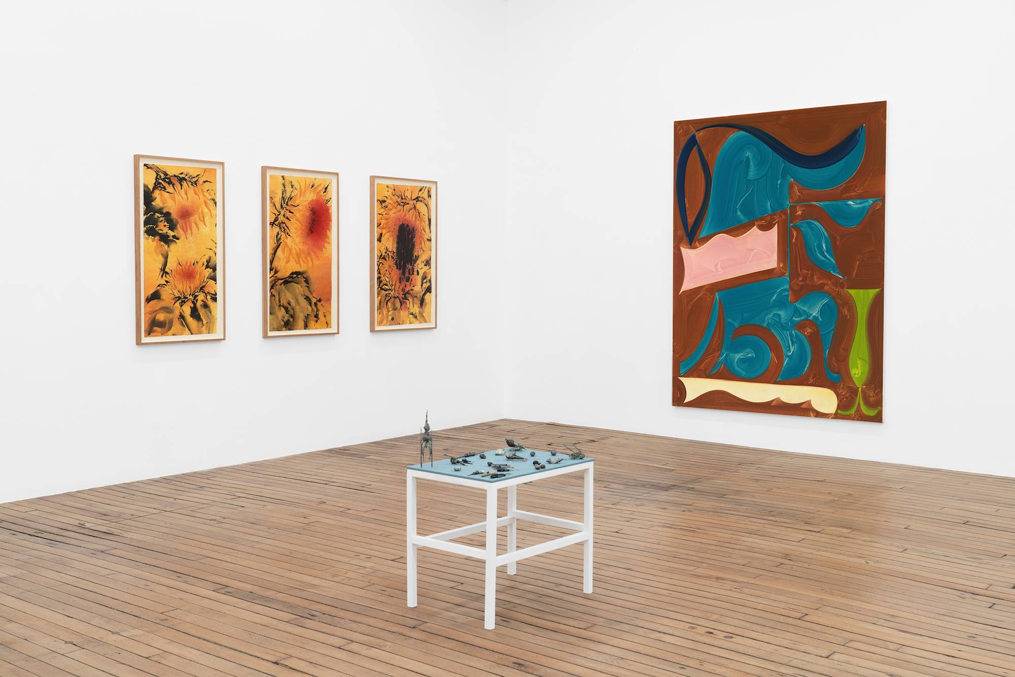 The corner of an art gallery displays a collection of paintings and a small, simple table with blue fabric top and sculptural elements.