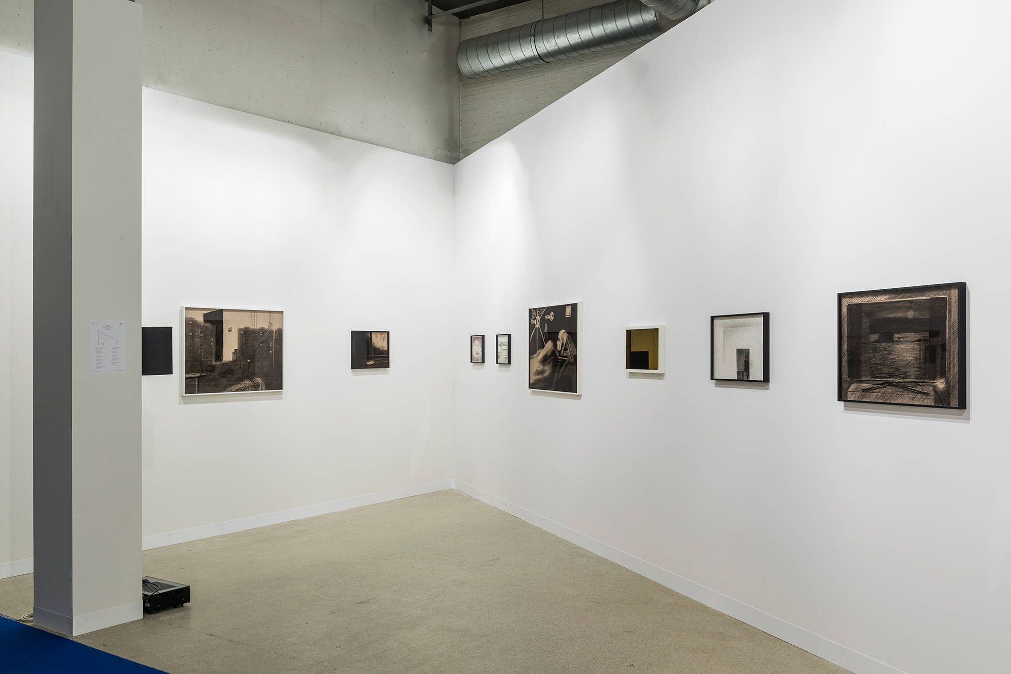Installation view of an art fair booth with an angled back wall, looking towards the back corner of the booth, framed drawings hung in a row on the two walls.