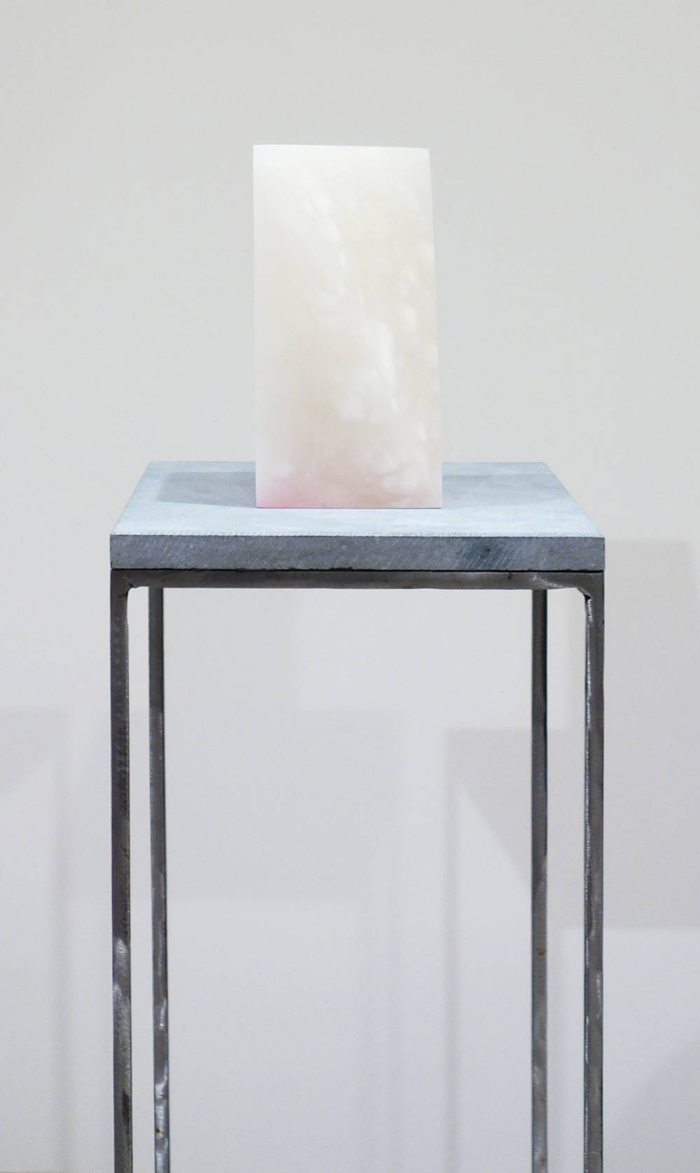 Atop a blue stone and steel plinth sits a rectangular block of white alabaster. There is a triangular piece of the alabaster cut from the block, and on the bottom is a gradient of pink acrylic paint.