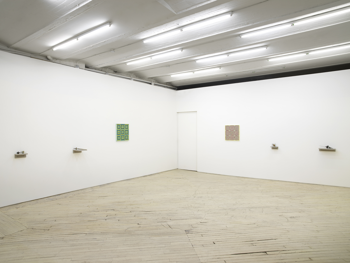 In a gallery space, two small abstract sculptures are installed on small gray shelves. Next to them is a square painting depicting a grid pattern in a range of greens. To the right is a painting of identical size and a similar pattern motif in red and yellow. To the right are two small sculptures installed on gray shelves. 