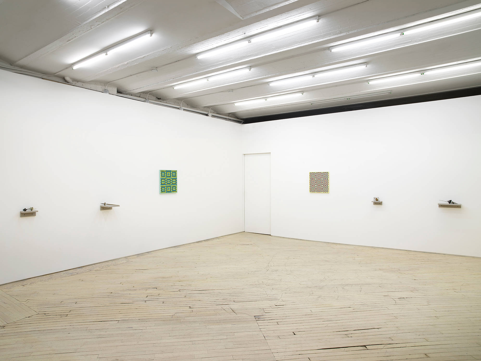 In a gallery space, two small abstract sculptures are installed on small gray shelves. Next to them is a square painting depicting a grid pattern in a range of greens. To the right is a painting of identical size and a similar pattern motif in red and yellow. To the right are two small sculptures installed on gray shelves.