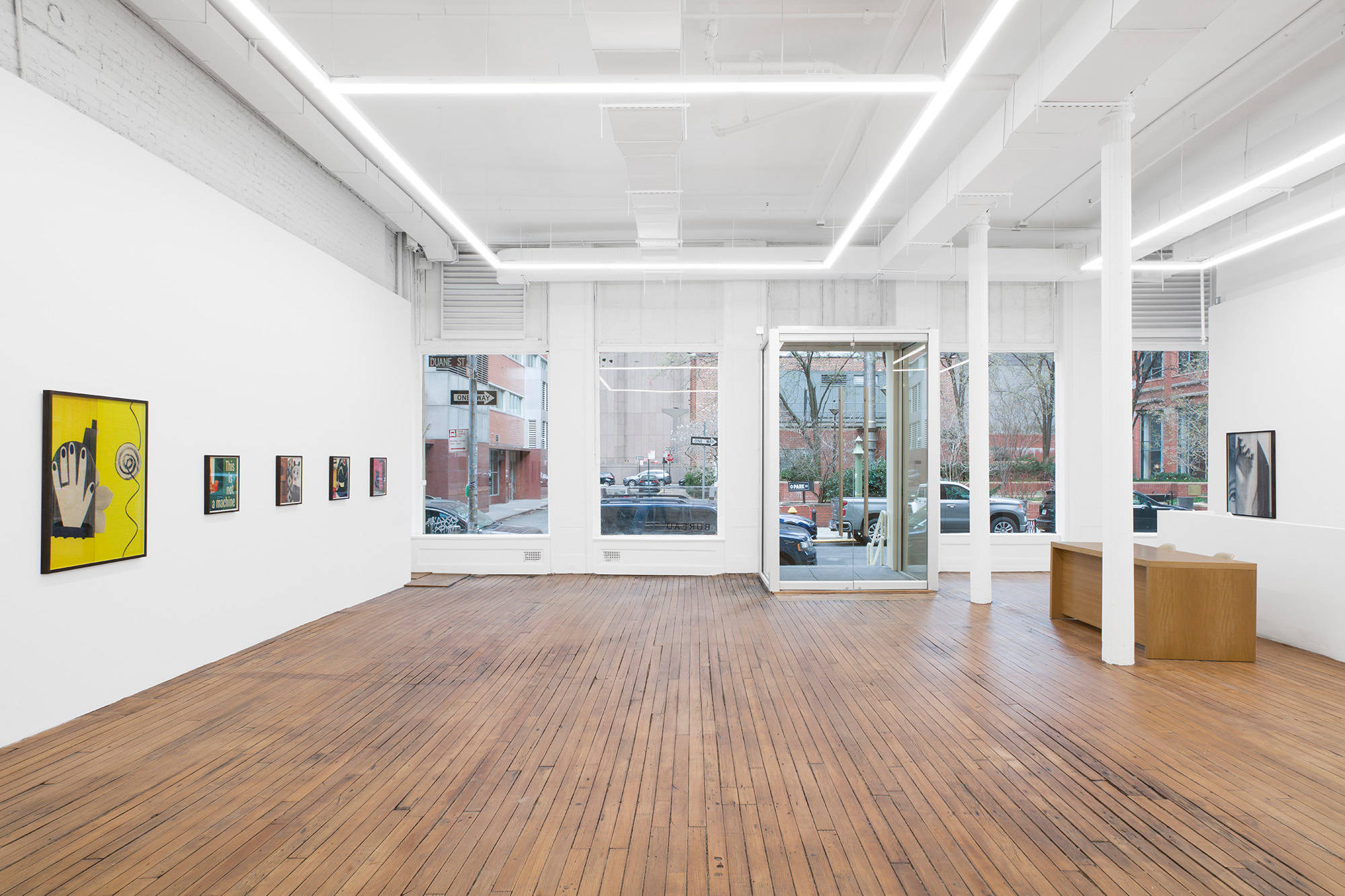An image of a large gallery space taken from the back of the gallery, facing the front doors. Four small and one large colorful artworks are hung on the left. One artwork can be seen hung on the right.