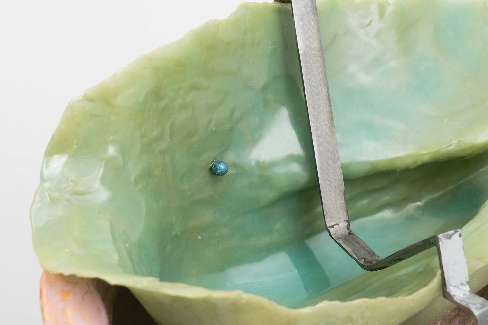 Detail of an Em Rooney sculpture featuring a stool wrapped in foil and mother of pearl, supporting a hinged steel mold of a flower, a pink fiberglass mold of an arm and a thin green silicone cast. A thin strip of bent steel is draped over the sculpture.