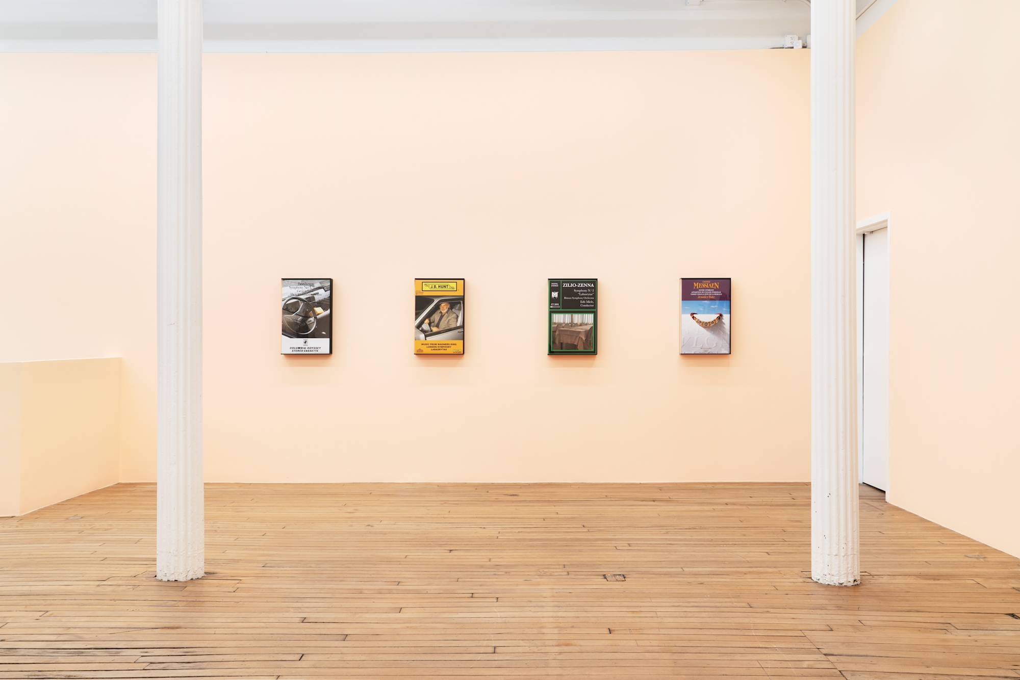 Four large, fake audio cassette cases featuring various artworks are displayed on a peach-colored wall in a gallery with wooden floors.