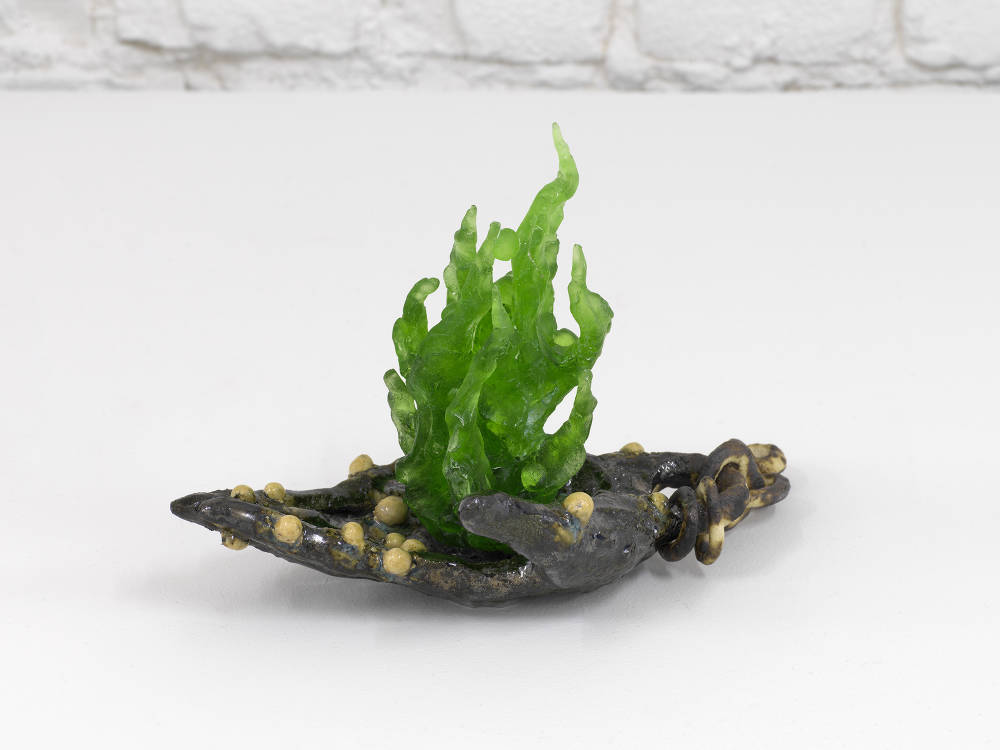 A small ceramic sculpture resembling an abstract human hold a cast of green glass in the shape of a flame. 
