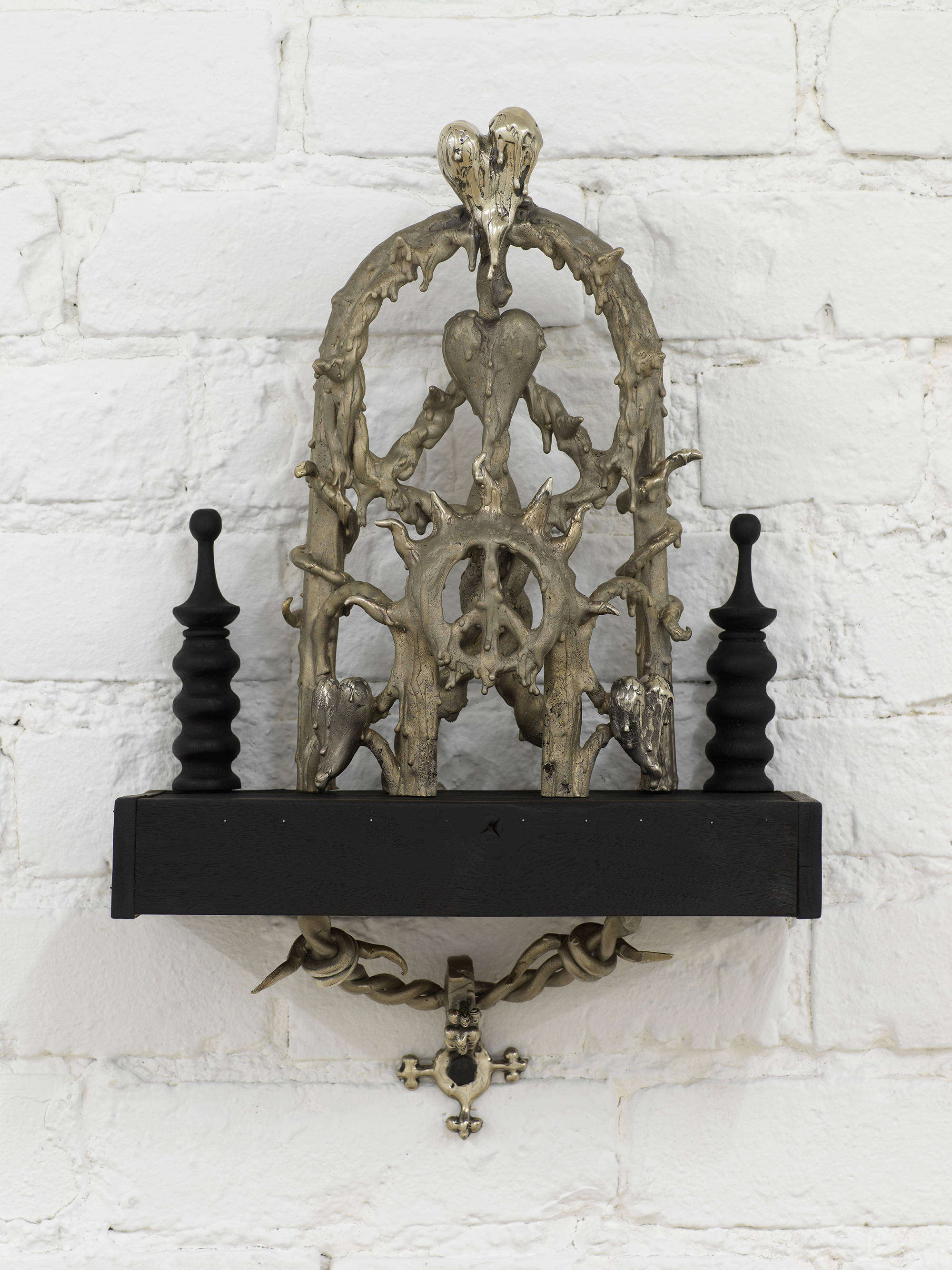 Detail view of Harvey's sculptural installation: an ornate cast bronze gothic style object sits on a black wooden shelf with turned wood decorate elements on left and right. The central bronze object features a rounded extended arch made up of concentric peace signs, melting hearts and wavy sun rays and spiky vine forms . Underneath the black shelf is another bronze cast of twisted barbed wire or vine, and a 4 pointed anchor which is bolted to the wall.