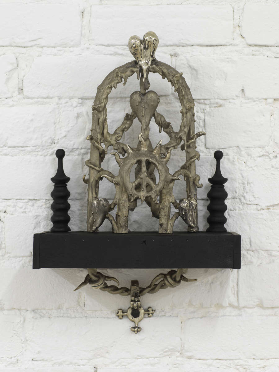 Detail view of Harvey's sculptural installation:  an ornate cast bronze gothic style  object sits on a black wooden shelf with turned wood decorate elements on left and right. The central bronze object features a rounded extended arch made up of concentric peace signs, melting hearts and wavy sun rays and spiky vine forms . Underneath the black shelf is another bronze cast of twisted barbed wire or vine, and a 4 pointed anchor which is bolted to the wall.