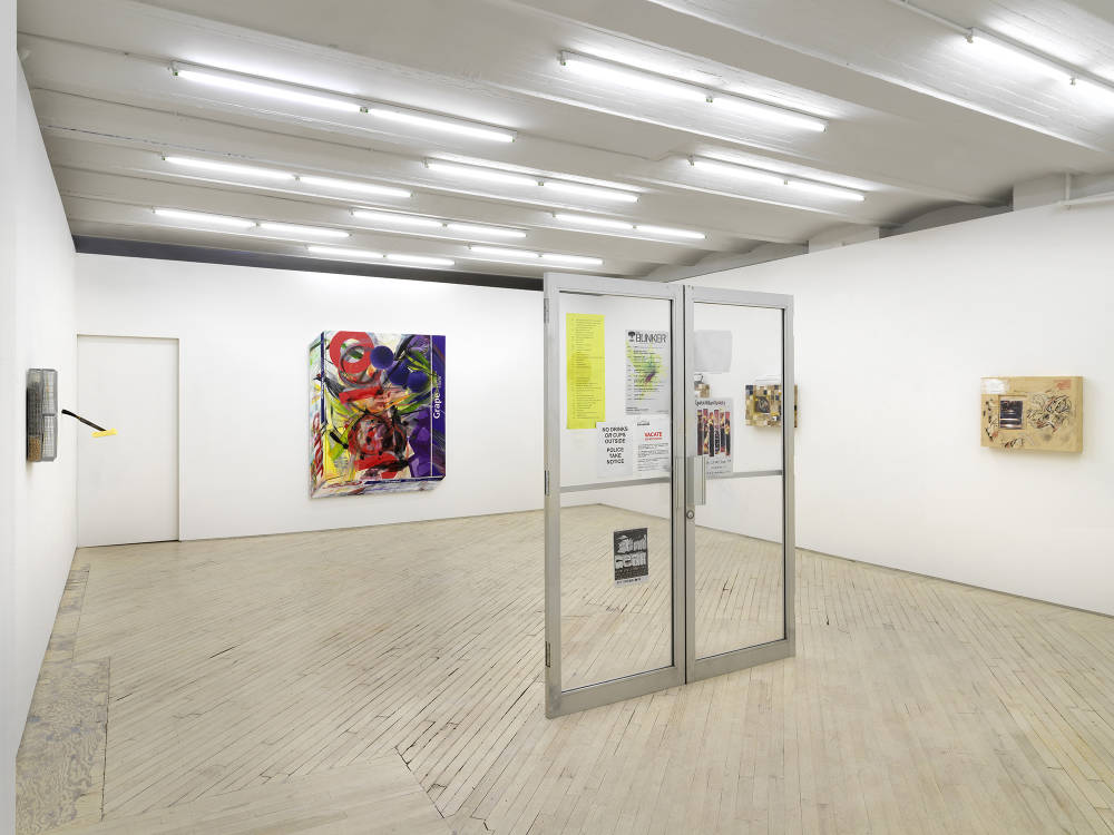 In a gallery space, a wall hanging metal sculpture next to another wall sculpture resembling a knife protruding from the wall with a stick of butter attached to the blade. In the center of the room is a freestanding glass doorway with poster advertisements attached to the glass. In the background is large painting resembling a box of Jello with abstract marks paint on its surface. To the right is a wall sculpture resembling the marks of a cave wall with a photograph adhered to it.