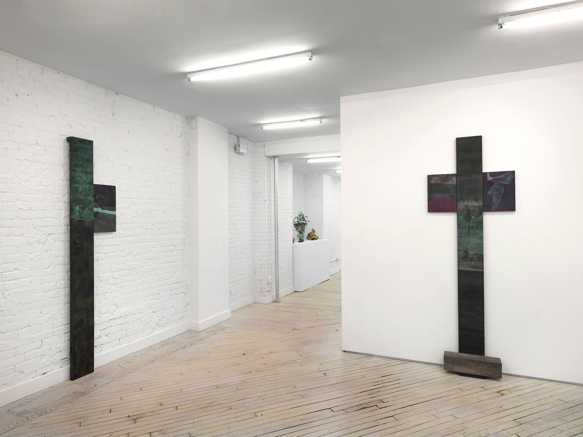Two vertical sculptures and paintings rendered in a dark palette leaning against gallery walls. In the distance a group of ceramics are placed on top of white pedestal.