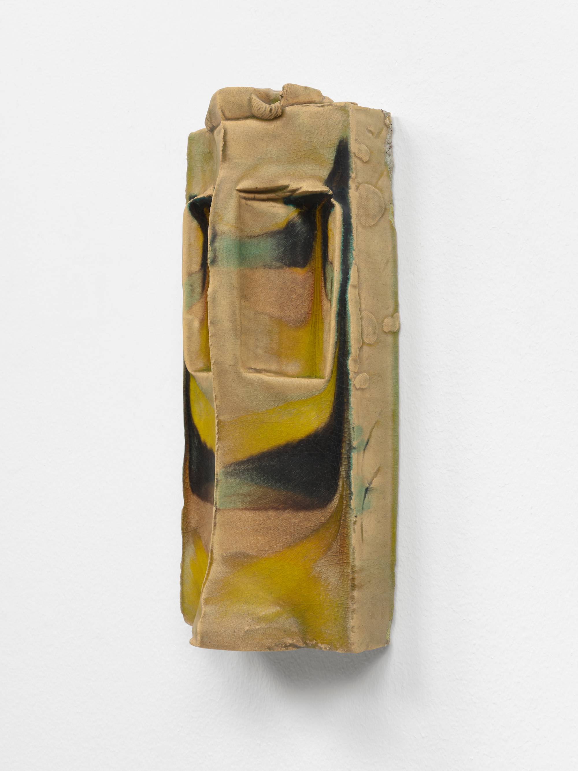 Three quarter view of a symmetrical, wall-mounted, ceramic sculpture composed of two narrow, vertical slopes that form a peak that runs parallel to the wall. In the top half of the work a shallow pocket has been carved out of each slope and the crown has been embellished with a small, curled ornament. The sculpture is glazed in hues of brown, green, and yellow.